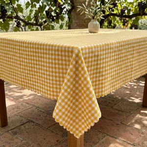 May include: A rectangular tablecloth featuring a yellow and white gingham check pattern with a scalloped edge. The fabric is draped over a wooden table, and a small, light-coloured ceramic vase containing a green plant sits on the surface.