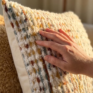 Puede incluir: Un coj&iacute;n cuadrado con una superficie texturizada y peluda sobre una base de color crema. La tela est&aacute; decorada con rayas verticales de peque&ntilde;os mechones en relieve en tonos apagados de amarillo mostaza, naranja quemado y azul polvoriento.