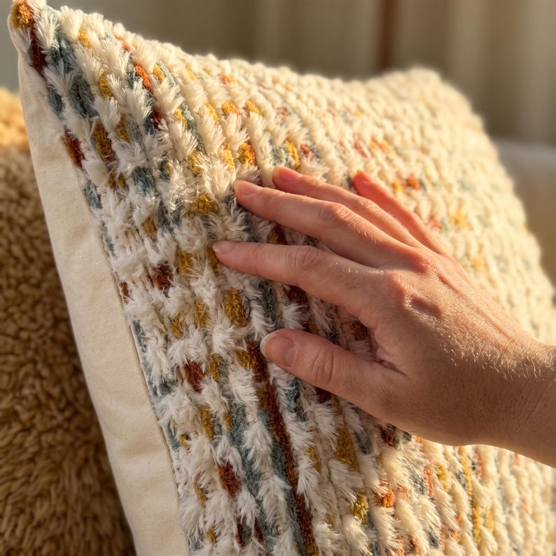 Puede incluir: Un coj&iacute;n cuadrado con una superficie texturizada y peluda sobre una base de color crema. La tela est&aacute; decorada con rayas verticales de peque&ntilde;os mechones en relieve en tonos apagados de amarillo mostaza, naranja quemado y azul polvoriento.