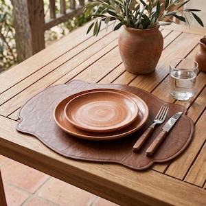May include: A brown leather placemat with a scalloped edge, featuring two stacked round terracotta-colored ceramic plates, a stainless steel fork with a wooden handle, and a stainless steel knife with a wooden handle on the right side.