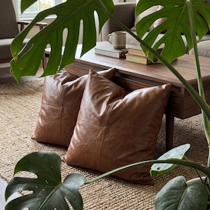 May include: Two square brown faux leather scatter cushions featuring a visible cross-stitched seam pattern on the front surface. The cushions have a smooth, matte texture and are positioned on a woven rug in front of a wooden coffee table.