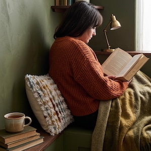 Puede incluir: Un coj&iacute;n decorativo de color crema con textura y rayas verticales de tela deshilachada en tonos &oacute;xido, azul y blanco. El coj&iacute;n est&aacute; colocado junto a una pila de libros, una taza de cer&aacute;mica y una manta tejida de color verde oliva suave.
