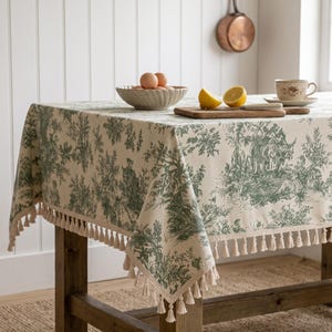 Könnte beinhalten: Eine cremefarbene Tischdecke mit grünem Toile-de-Jouy-Muster und dekorativen Fransen an den Rändern. Der Stoff liegt auf einem Holztisch, auf dem sich eine Schale mit Eiern, ein Holzschneidebrett mit Zitronenhälften und eine Tasse befinden.