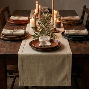 May include: A rustic wooden dining table set for four with a beige linen table runner. Each place setting features a dark brown plate, a beige linen napkin tied with twine, and a centerpiece of lit taper candles and green olive branches.