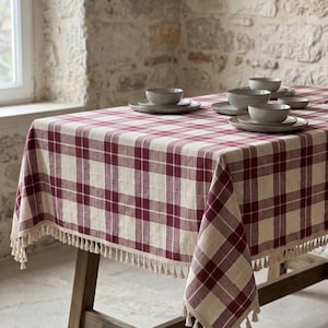 May include: A rectangular tablecloth featuring a burgundy and cream plaid pattern with a fringe trim along the edges. The fabric is draped over a wooden table, which is set with several grey ceramic bowls and plates arranged on the surface.