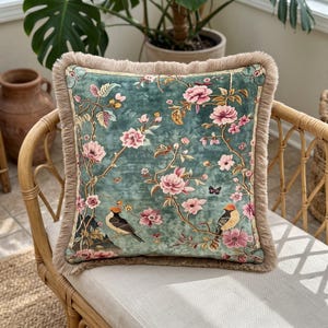 Puede incluir: Un cojín cuadrado de tela de terciopelo color verde azulado con un estampado floral de flores rosas, ramas y dos pájaros pequeños. El cojín está rematado con un fleco grueso de color marrón claro en los cuatro bordes del cojín.