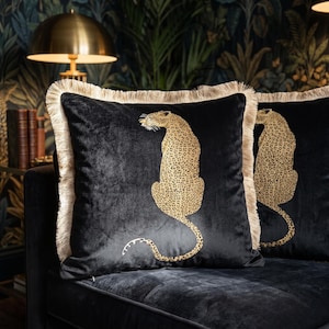 May include: A square black velvet throw pillow featuring a gold and black leopard print graphic in the center. The pillow is finished with a long, light beige fringe trim along all four edges, creating a textured border around the dark fabric.