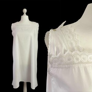 May include: A white nightdress with a square neckline and lace detailing. The sleeveless garment is displayed on a mannequin against a black backdrop. The lace trim is visible on the neckline and shoulders.