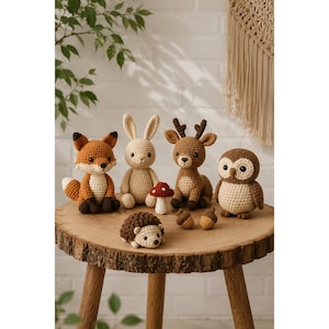 May include: A collection of six crocheted stuffed animals including a fox, a rabbit, a deer, an owl, and a hedgehog, accompanied by a small red and white mushroom and two brown acorns, all arranged together on a round wooden surface.