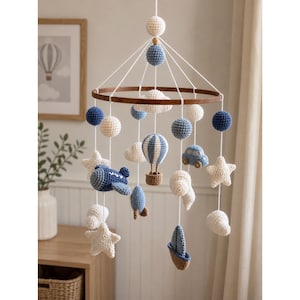 Può includere: Una giostrina sospesa con figure all'uncinetto nei toni del blu, bianco e marrone. La giostrina include una mongolfiera, un aereo, una barca a vela, un'auto, un razzo, stelle e nuvole sospese a una cornice circolare in legno con fili bianchi.
