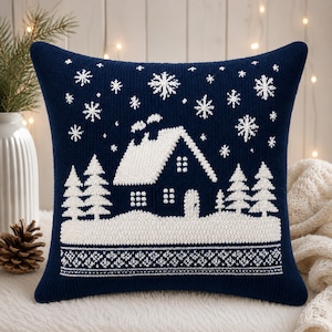 May include: A square navy blue knit throw pillow featuring a white winter scene with a house, evergreen trees, and falling snowflakes. The bottom section displays a detailed white geometric pattern, and the entire design is knitted into the fabric.