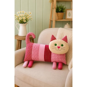 May include: A cat-shaped decorative pillow featuring a patchwork body with red and white gingham, pink stripes, and solid red fabric. The pillow has a cream-colored face with green button eyes, a pink nose, and embroidered whiskers and ears.