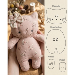 Puede incluir: Un peluche de gato rosa claro con un delicado estampado floral y un lazo de tela a juego en la oreja. Junto al juguete, un diagrama muestra las piezas del patrón de costura etiquetadas como Plasmalle x 2, Fielerhautrag x 2, Armnel x 4 y Pofden.