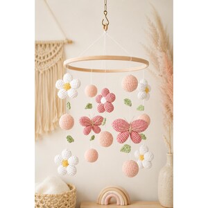 May include: A hanging mobile featuring a circular wooden frame with dangling crochet elements including white and pink flowers, pink butterflies, small green leaves, and solid pink spheres suspended by thin white strings from the central hoop.