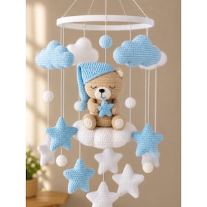 May include: A crochet baby mobile featuring a central tan bear wearing a blue nightcap and holding a blue star. The mobile includes white and blue crocheted clouds, stars, and small spheres suspended from a white circular frame by white strings.