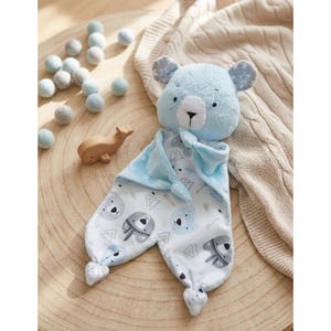 Peut inclure: Une couverture de sécurité en peluche bleu clair en forme d'ours, dotée d'une tête douce aux traits brodés et d'oreilles à motifs d'étoiles. La couverture présente un fond blanc avec un motif répétitif de têtes d'ours, de flèches et de triangles.