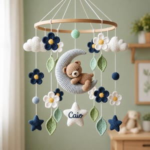 Pode incluir: Um móbile de crochê para bebê com uma lua crescente azul-clara central com um ursinho de pelúcia marrom, cercado por flores brancas e azul-escuras, folhas verdes, nuvens brancas e estrelas. Uma estrela branca na base tem o nome Caio bordado.