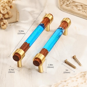 May include: Two cabinet pulls featuring a cylindrical wood body with a bright blue resin insert, accented by brushed gold metal end caps and posts. The pulls measure 5.39 inches in length, 1.38 inches in height, and 0.75 inches in diameter, with 1-inch screws.