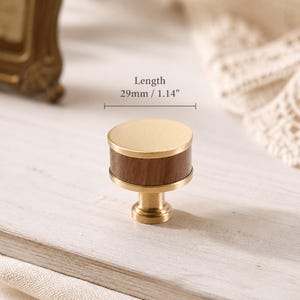 Peut inclure: Un bouton de meuble cylindrique composé d'une bande centrale en bois brun foncé prise entre deux disques en métal doré brossé. Le bouton possède une base en métal doré assortie et une longueur totale de 29 mm indiquée par le texte sur l'image.