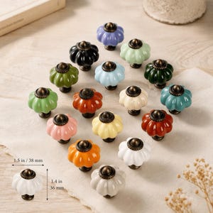 Op de afbeelding: Een collectie van vijftien pompoenvormige keramische kastknoppen in diverse kleuren, waaronder wit, zwart, groen, blauw, geel, oranje, roze en rood. Elke knop heeft een bronskleurige metalen voet en top, met een breedte van 38 mm en hoogte van 36 mm.