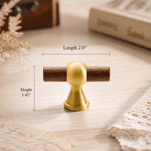 May include: A T-shaped cabinet knob featuring a horizontal dark brown wooden cylinder centered with a brushed gold metal sphere and a conical base. The knob has a length of 2.0 inches and a height of 1.42 inches as indicated by the text labels.