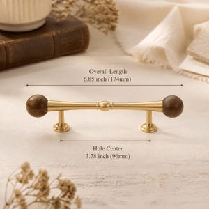 May include: A cabinet pull featuring a central gold-toned metal bar with a small sphere in the middle and dark brown spherical ends. The pull has an overall length of 174mm and a hole centre distance of 96mm as indicated by the text in the image.