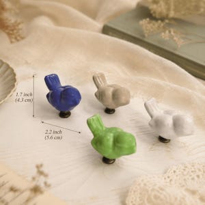 May include: Four bird-shaped ceramic cabinet knobs in blue, beige, green, and white. The knobs measure 1.7 inches in height and 2.2 inches in width. Each knob features a rounded bird silhouette with a small tail and a dark metal mounting base.