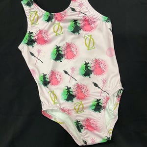 May include: A light pink one-piece swimsuit with a pattern of witches, brooms, and the letter "D". The design includes pink, green, and black elements. The swimsuit has a scoop neck and a high-cut leg.