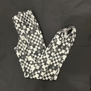 May include: Black and white checkered pattern pants. The pants are folded and laid out on a black surface. The pattern is a repeating grid of black and white squares.