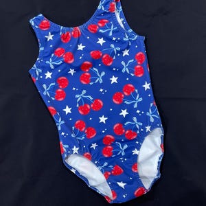 May include: A royal blue leotard featuring a repeating pattern of red cherries with light blue and white striped stems and bows, interspersed with small white five-pointed stars. The garment has a scoop neckline and a sleeveless cut.