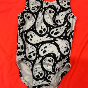 May include: A sleeveless bodysuit with a black and white ghost pattern. The design features cartoon ghosts on a white background, with black outlines and details. The garment is likely for a child, with elasticated leg openings.