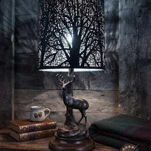 May include: A table lamp featuring a dark brown deer figurine as the base, standing on a circular wooden pedestal. The cylindrical lampshade is black with a laser-cut silhouette pattern of bare tree branches, allowing light to shine through.