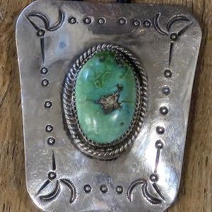 May include: A bolo tie featuring a rectangular silver-toned metal plate with rounded corners and stamped decorative markings. A central oval-shaped green turquoise stone with dark matrix veins is set in a twisted rope border on a black cord.