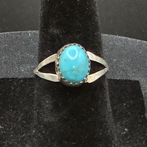 Op de afbeelding: Een zilveren ring met een centrale ovaalvormige turkoois edelsteen met zichtbare donkere matrixaders. De band is gespleten in twee dunne, gepolijste zilveren armen die samenkomen bij de zetting, die een decoratieve geschulpte rand heeft.