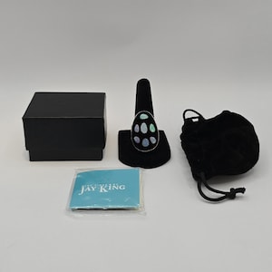 May include: A silver ring featuring an oval face with six inlaid iridescent opal stones, displayed on a black stand. The set includes a black square box, a black drawstring pouch, and a small blue card with the text Mine Finds by Jay King.