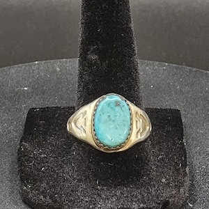 May include: A silver-toned ring featuring an oval-shaped turquoise stone set in a serrated bezel. The band displays engraved geometric patterns on both sides of the central stone, showcasing a polished metal finish and a vibrant blue-green gemstone.