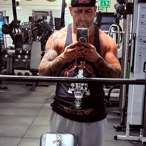 May include: A black tank top featuring a graphic of a bodybuilder and the text GOLD'S GYM in red and white lettering, paired with light gray athletic shorts and a black baseball cap worn backwards, captured in a mirror reflection.