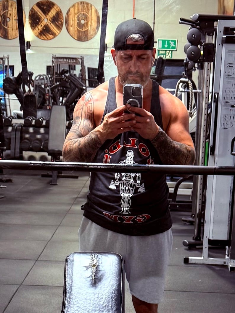 May include: A black tank top featuring a graphic of a bodybuilder and the text GOLD'S GYM in red and white lettering, paired with light gray athletic shorts and a black baseball cap worn backwards, captured in a mirror reflection.