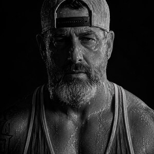 May include: A black and white portrait of a person wearing a baseball cap and a tank top. The individual has a full beard and is covered in visible moisture droplets, with a dark, high-contrast aesthetic emphasizing the texture of the skin and fabric.