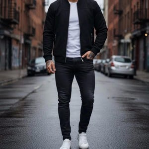 May include: A black bomber jacket with a front zipper, worn over a plain white t-shirt. The outfit includes slim-fit dark charcoal jeans and clean white sneakers. The jacket features ribbed cuffs and a collarless neckline, creating a layered look.