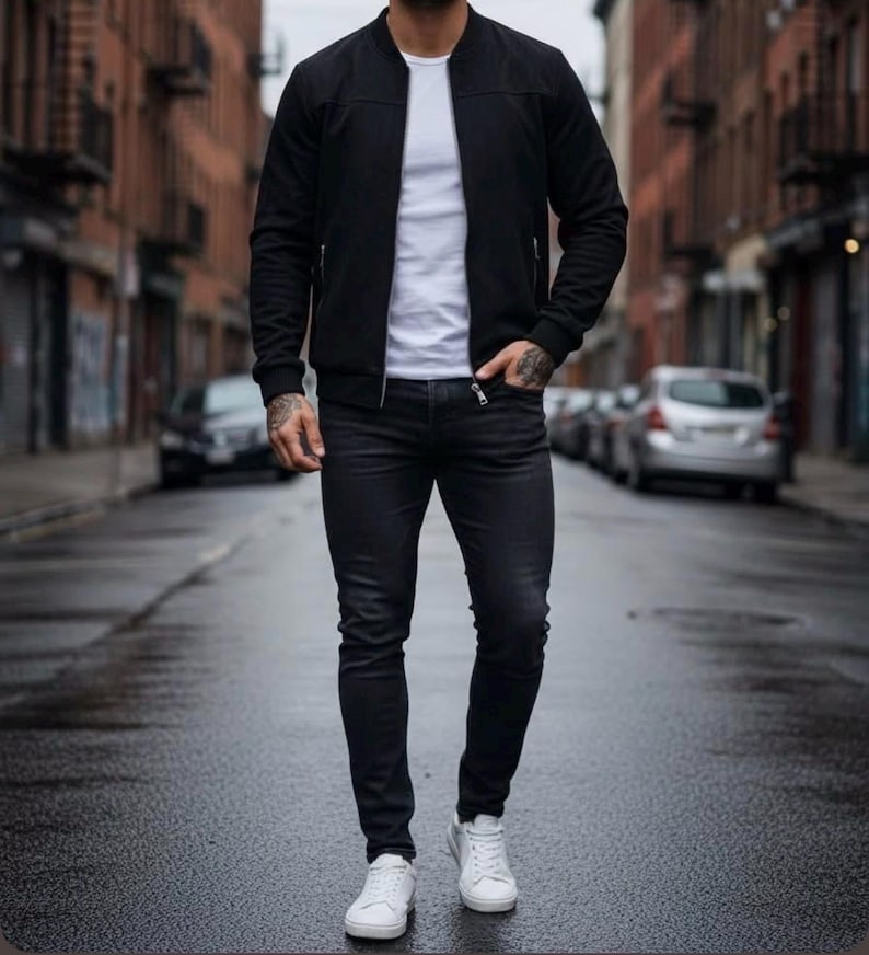 May include: A black bomber jacket with a front zipper, worn over a plain white t-shirt. The outfit includes slim-fit dark charcoal jeans and clean white sneakers. The jacket features ribbed cuffs and a collarless neckline, creating a layered look.