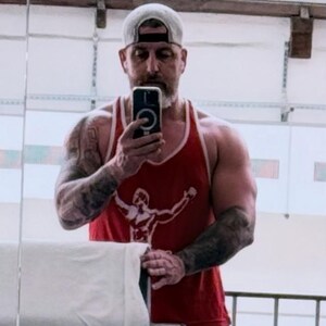 May include: A red sleeveless tank top featuring a white graphic print of a figure with arms raised. The garment has a scoop neckline and white trim along the armholes and neck. The fabric appears to be a lightweight cotton blend material.