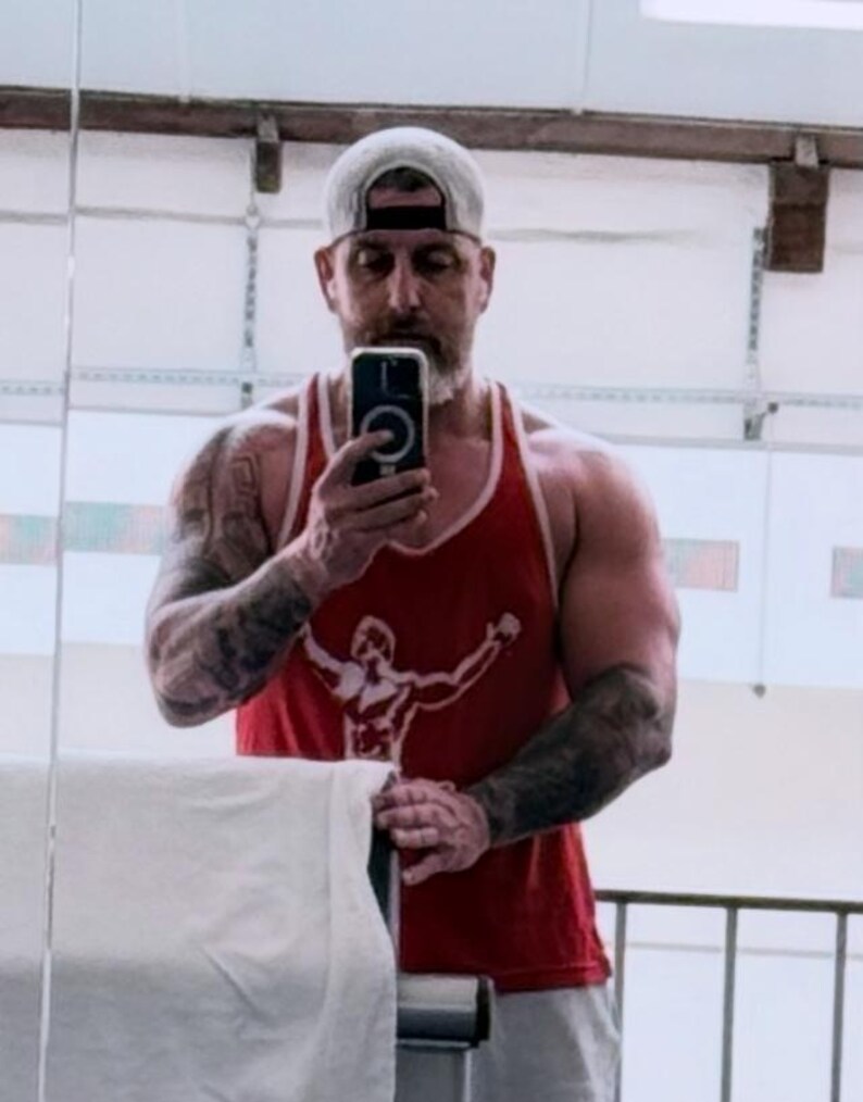 May include: A red sleeveless tank top featuring a white graphic print of a figure with arms raised. The garment has a scoop neckline and white trim along the armholes and neck. The fabric appears to be a lightweight cotton blend material.