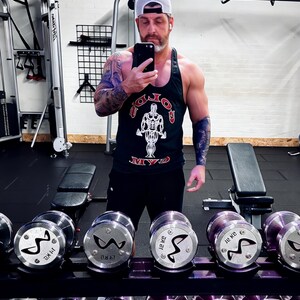 May include: A black tank top with a white and red Gold's Gym logo graphic, paired with black shorts and a white baseball cap. The tank top features a central illustration of a bodybuilder and the text Gold's Gym in a bold, arched font.