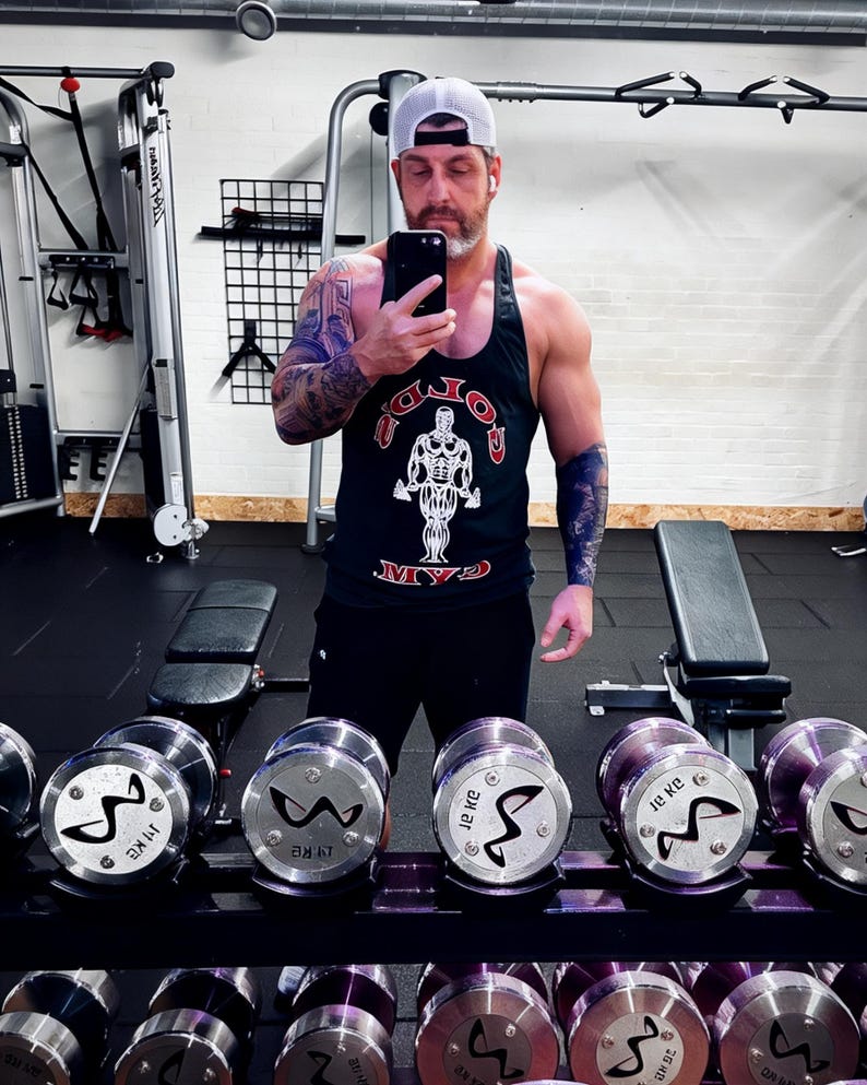 May include: A black tank top with a white and red Gold's Gym logo graphic, paired with black shorts and a white baseball cap. The tank top features a central illustration of a bodybuilder and the text Gold's Gym in a bold, arched font.