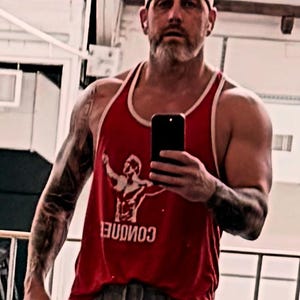 May include: A red sleeveless tank top with white trim around the neck and arm openings. The front of the shirt features a white graphic of a person with arms raised and the word CONQUER printed in white capital letters below the graphic.