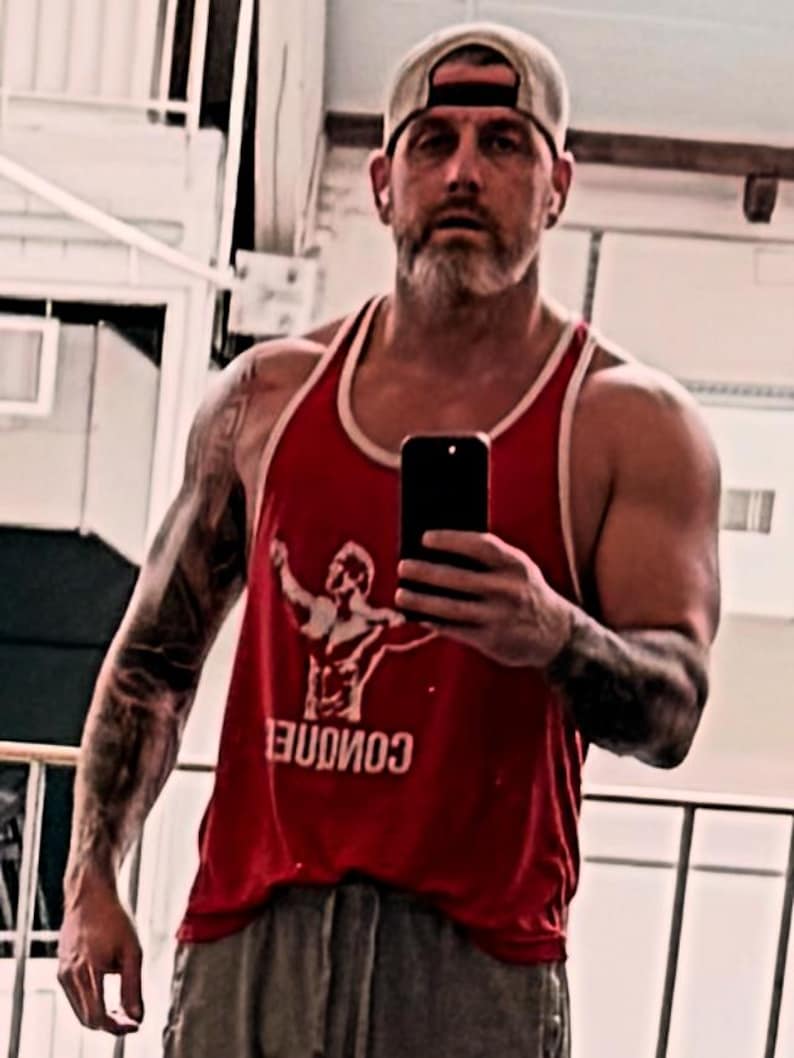 May include: A red sleeveless tank top with white trim around the neck and arm openings. The front of the shirt features a white graphic of a person with arms raised and the word CONQUER printed in white capital letters below the graphic.