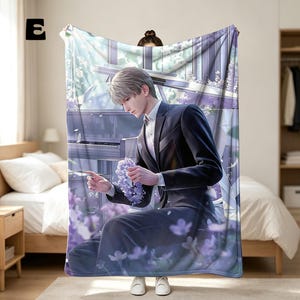 May include: A rectangular throw blanket featuring a printed illustration of a person with short light hair wearing a dark suit and white shirt, holding a cluster of purple flowers. A grand piano is visible in the background with purple floral accents.