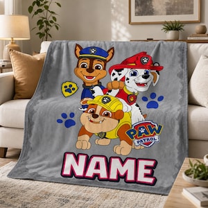May include: A gray fleece blanket featuring Paw Patrol characters Chase, Marshall, and Rubble. The design includes blue paw prints, the Paw Patrol logo, and the word NAME in large white letters with a pink outline. The blanket has a soft texture.
