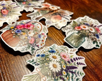 May include: A collection of die-cut stickers featuring various floral arrangements, including bouquets tied with twine, flowers in woven baskets, and blossoms in glass jars. The designs showcase daisies, cornflowers, and greenery in vibrant colors.